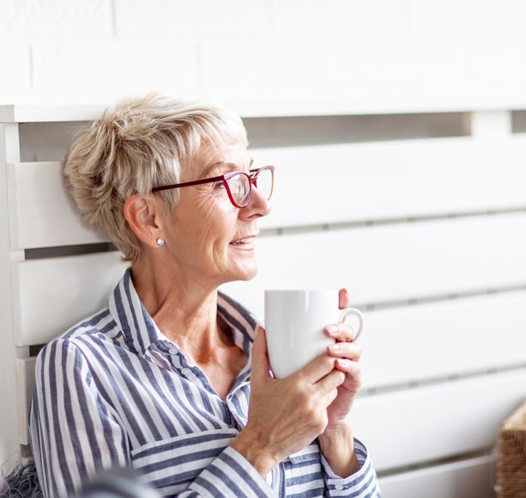 Mature good looking woman sitting in the room with mug on her hand and smile