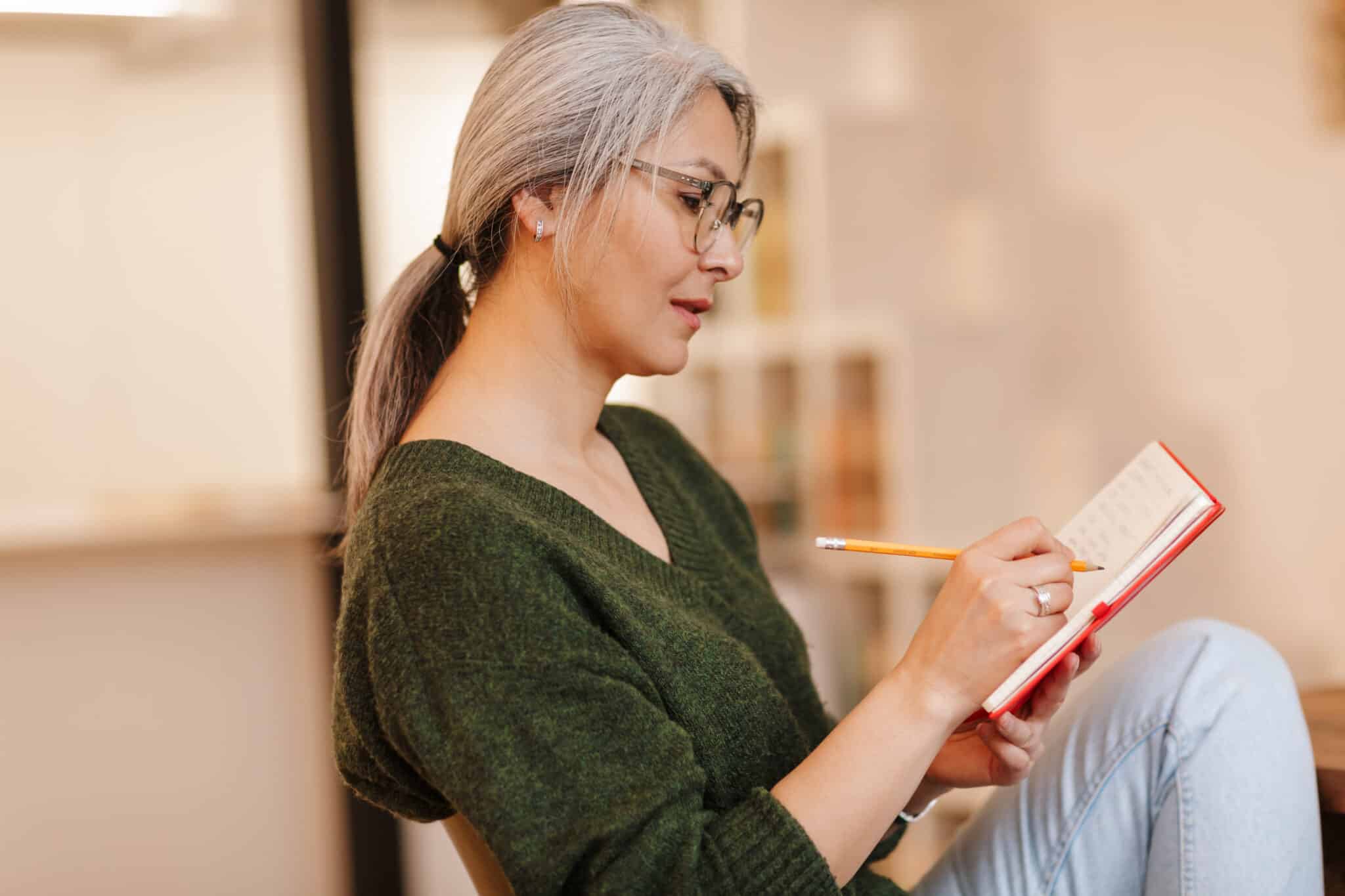 Photo of concentrated mature woman making notes in diary while sitting Photo of concentrated mature woman in eyeglasses making notes in diary while sitting at table in living room