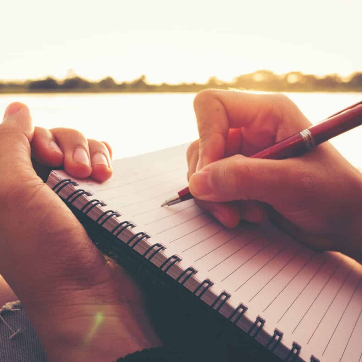 Close up hand of young woman with pen writing on notebook at riverside in the evening.