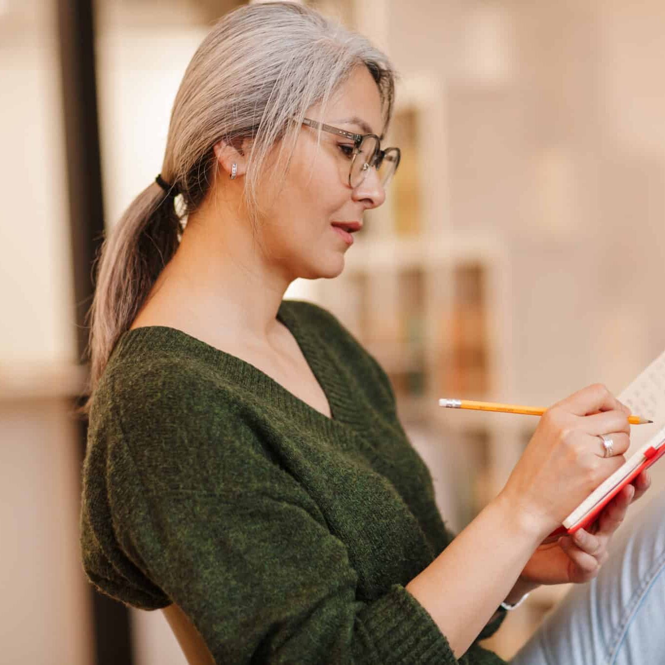 Photo of concentrated mature woman in eyeglasses making notes in diary while sitting at table in living room